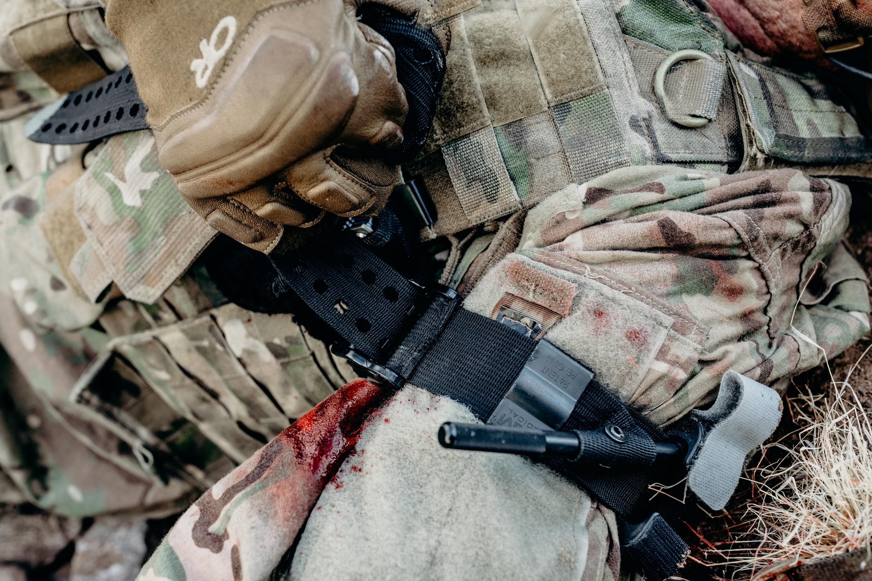 Eine mit einem beigen Handschuh bekleidete Hand hält das schwarze Extremity Tourniquet SAM® XT, befestigt an einem tarnfarbenen Rucksack mit Riemen und Schnallen.