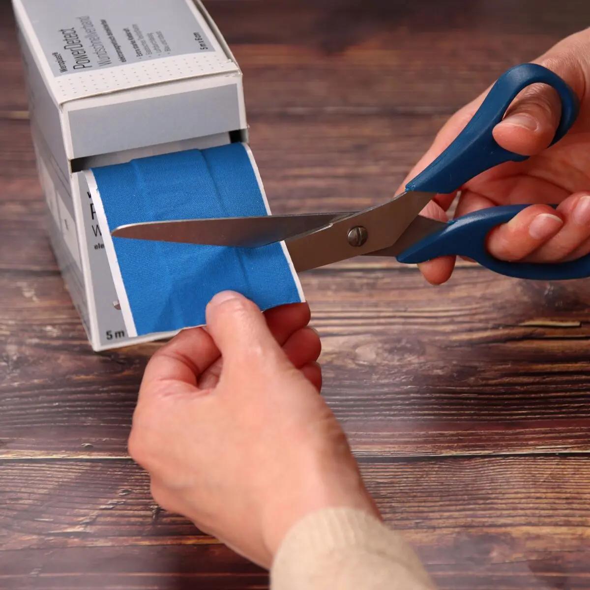 Zwei Hände schneiden mit blauen Scheren ein Stück vom Wundschnellverband Weroplast® PowerDetect, erkennbar an blauer Farbe und robustem Gewebe, ab.