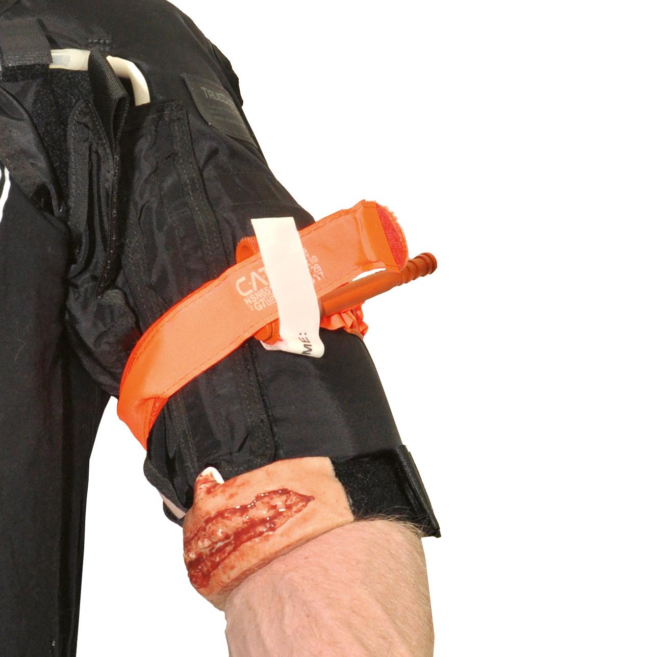 Schwarze taktische Weste mit orangefarbenem Notfallgurt und weißem Etikett, getragen über der Schulter, weißer Hintergrund.