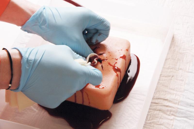 Zwei Hände in blauen Handschuhen üben an einem Erler-Zimmer Wound Packing Trainer aus strapazierfähigem Silikon mit realistisch blutender Schusswunde.
