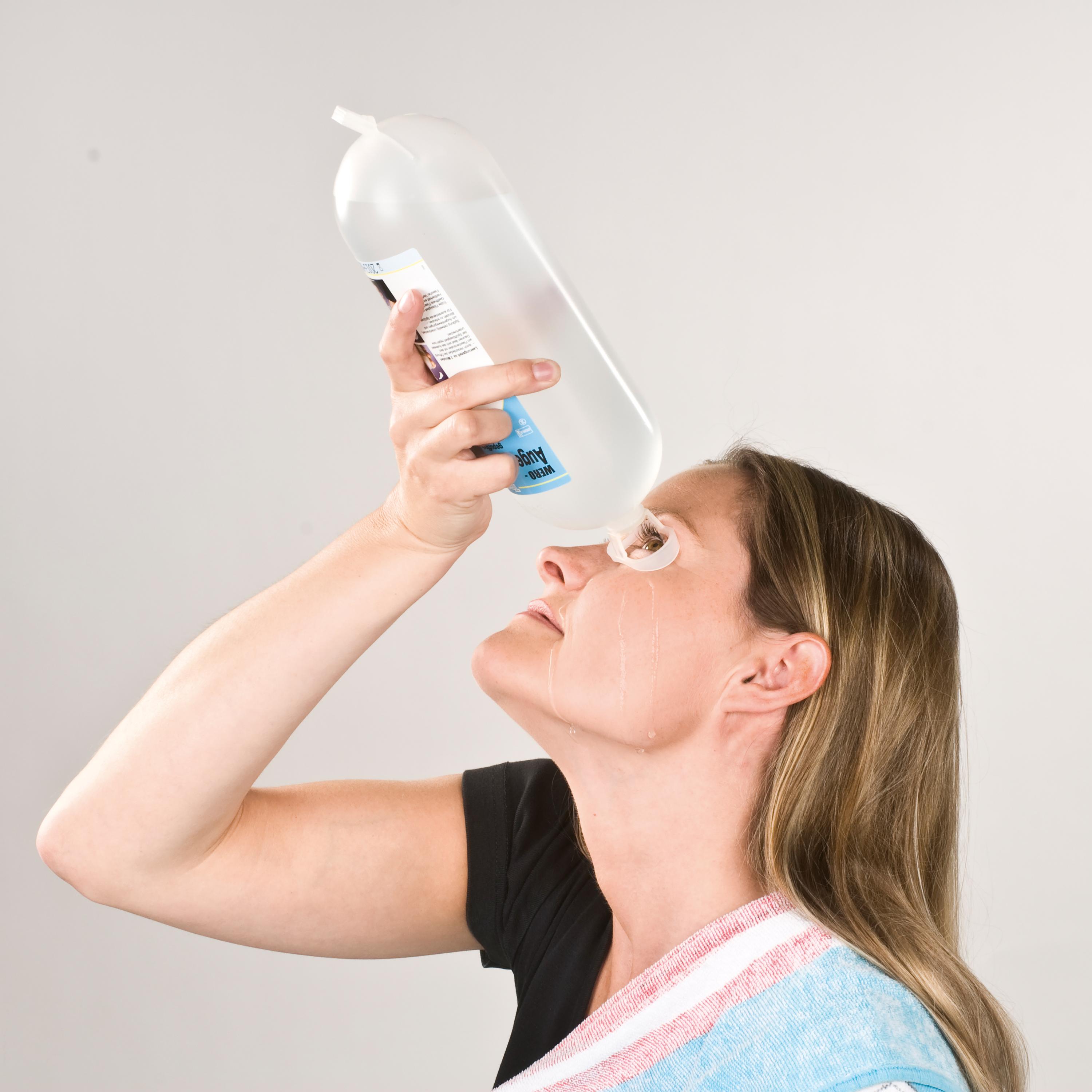 Eine Frau hält eine WERO Augenspülflasche Modell P 8 mit gepufferter, steriler Lösung über sich, während Flüssigkeit auf ihr Gesicht tropft.