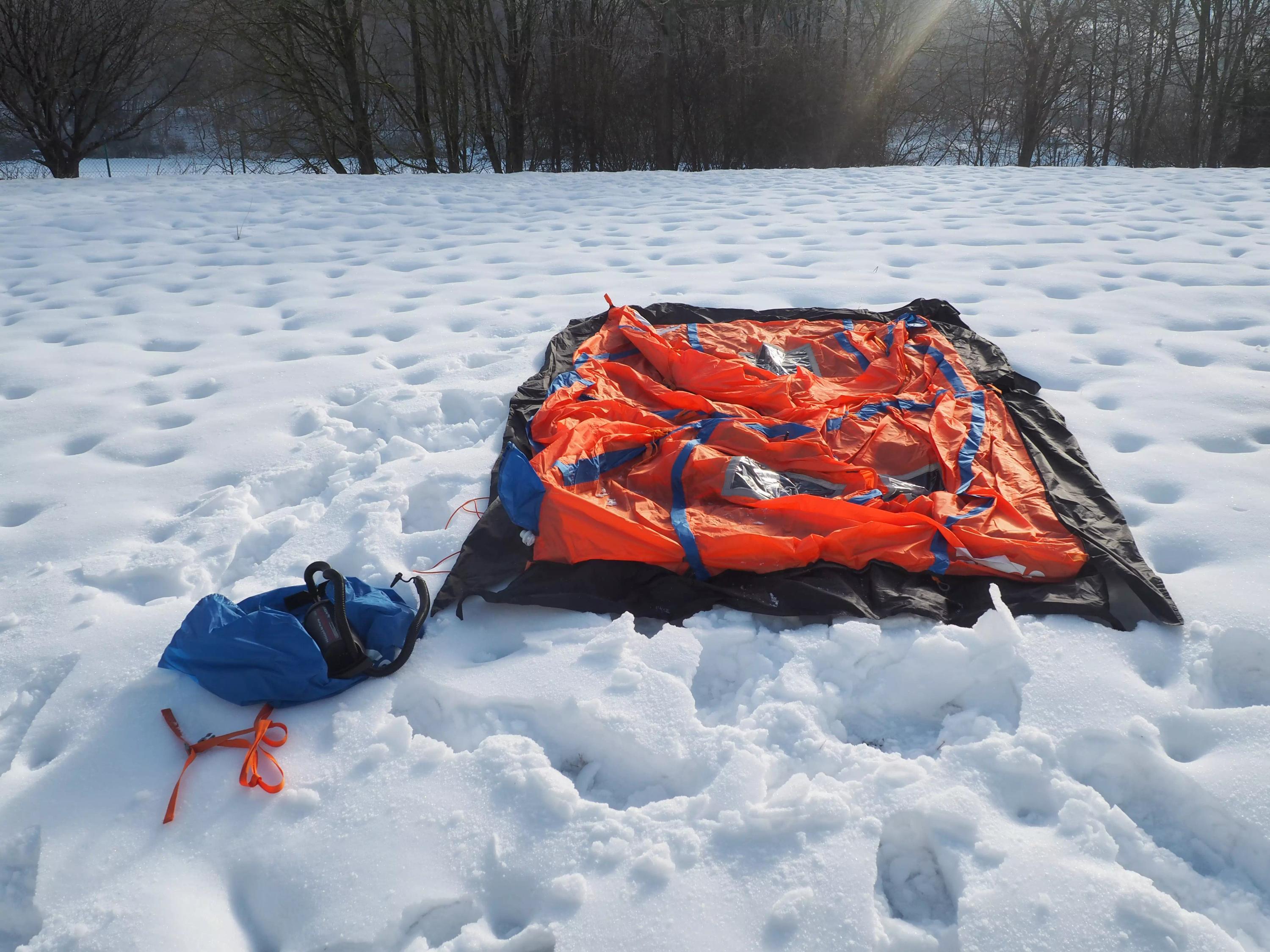 Ein orange-blaues RIGLOO Rescuer Rettungszelt steht halb eingeschneit mit sichtbar leichtem Gewicht und kompaktem Packmaß in verschneiter Landschaft.