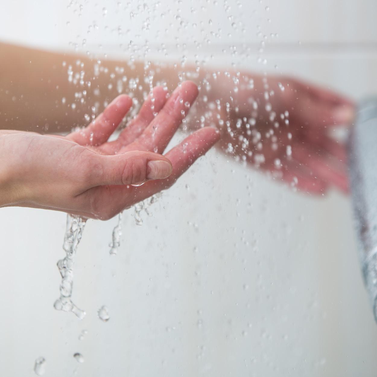 Eine Person befindet sich vermutlich unter der Dusche. Im Fokus befindet sich eine Hand, auf die das Wasser tropft.