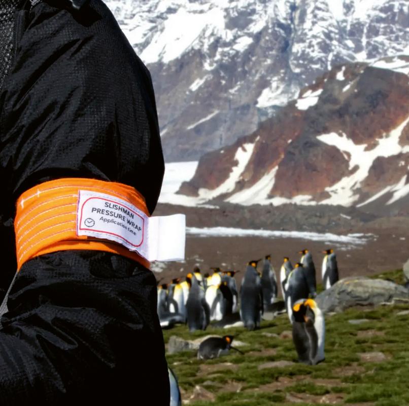Eine Person mit schwarzer Jacke trägt das orangefarbene Slishman Pressure Wrap als markantes Druckverband-Armband am Handgelenk.