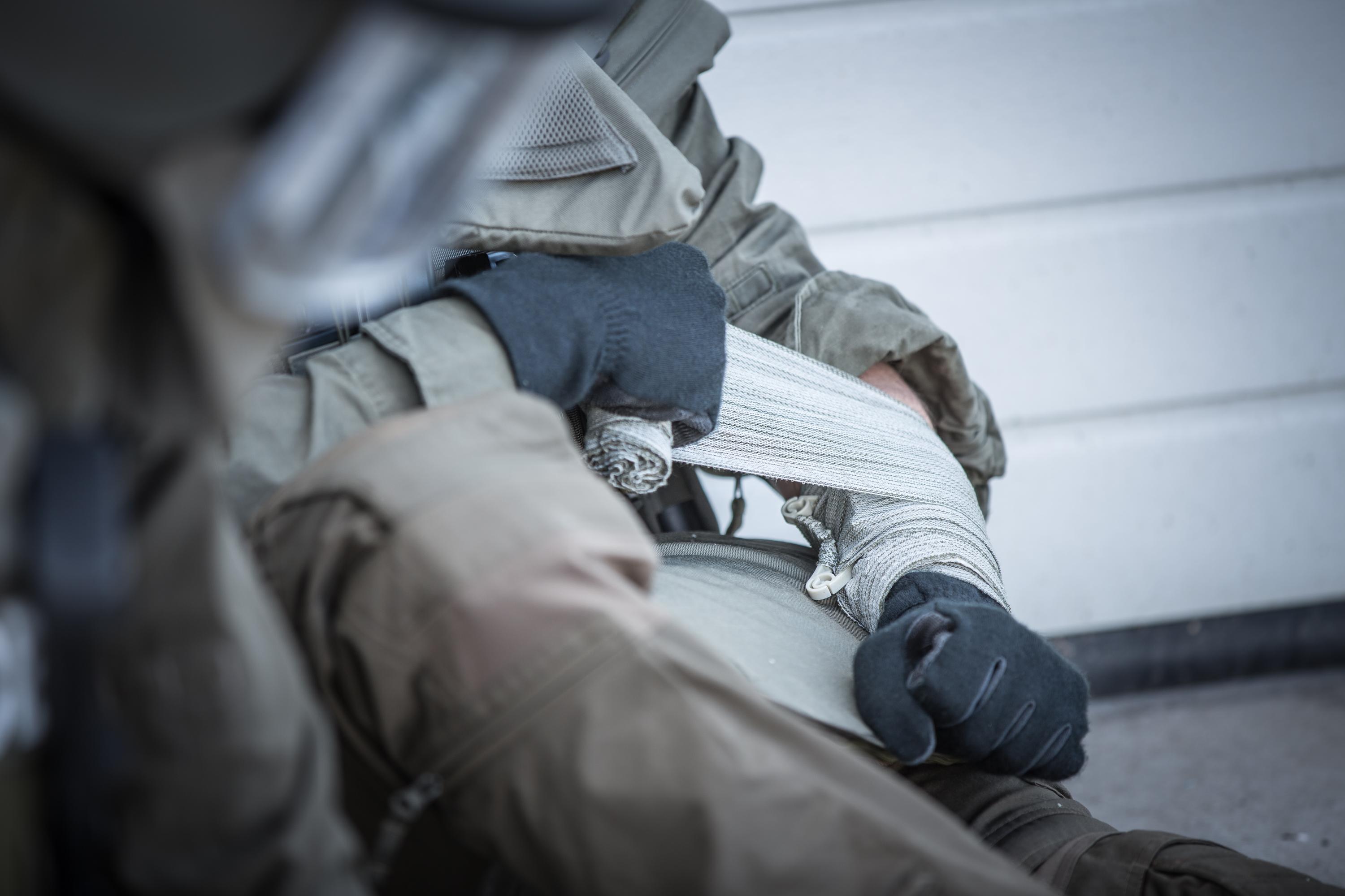 Eine Person in Tarnkleidung nutzt Die Emergency Bandage®, das ORIGINAL - Oliv, 10 cm als Notfallverband am Handgelenk mit sichtbarer weißer Bandage.