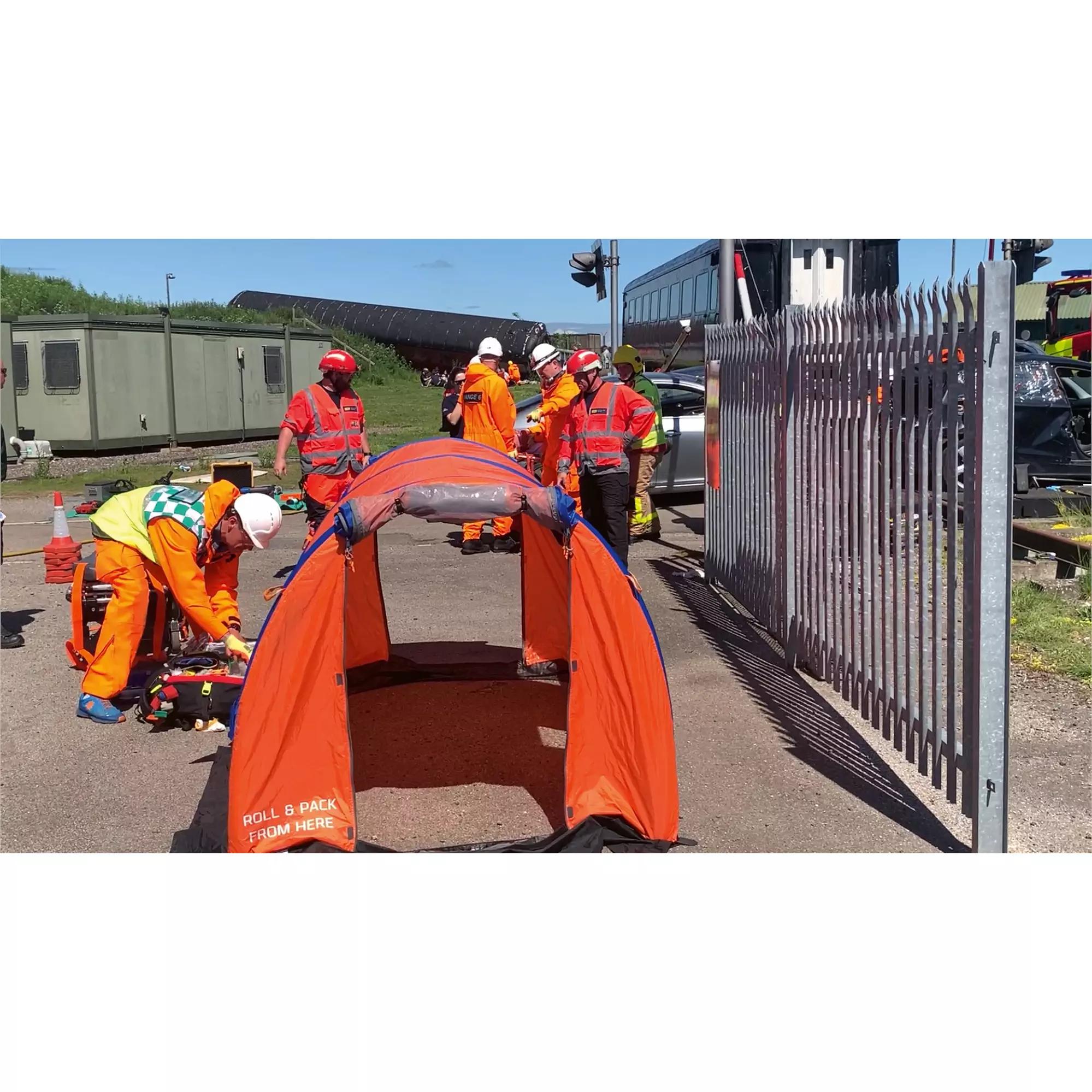 Mehrere Arbeiter in orangefarbener Kleidung errichten ein orangefarbenes RIGLOO Rescuer Rettungszelt mit eingebauten Ventilen neben Industrieanlagen.