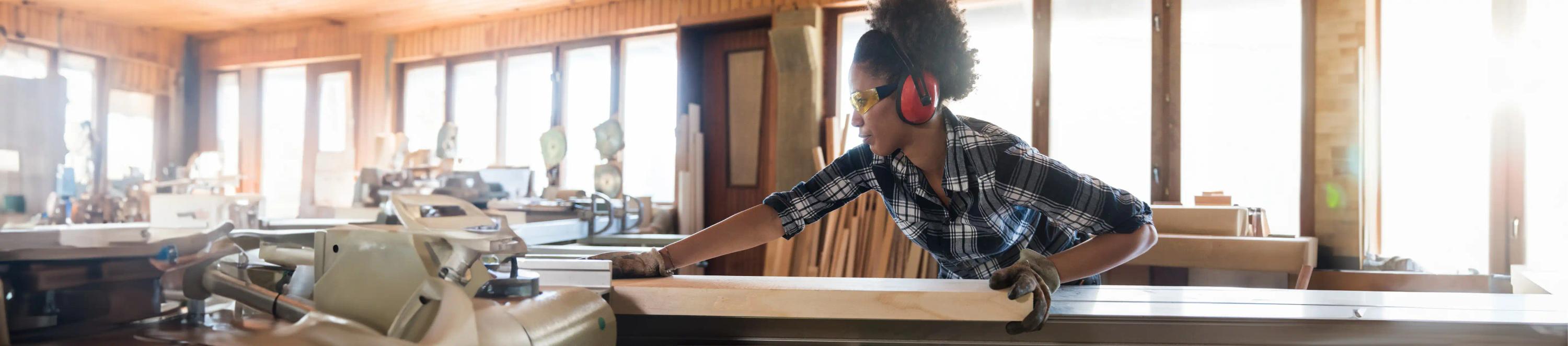 Eine Frau mit Schutzbrille und Handschuhen schneidet konzentriert ein Holzbrett mit einer elektrischen Säge auf einer Werkbank.
