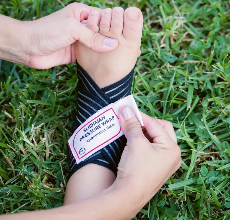 Zwei Hände legen einen Slishman Pressure Wrap - Schwarz mit weißem Etikett, elastischem Material und Lasso um einen Fuß auf grünem Gras an.