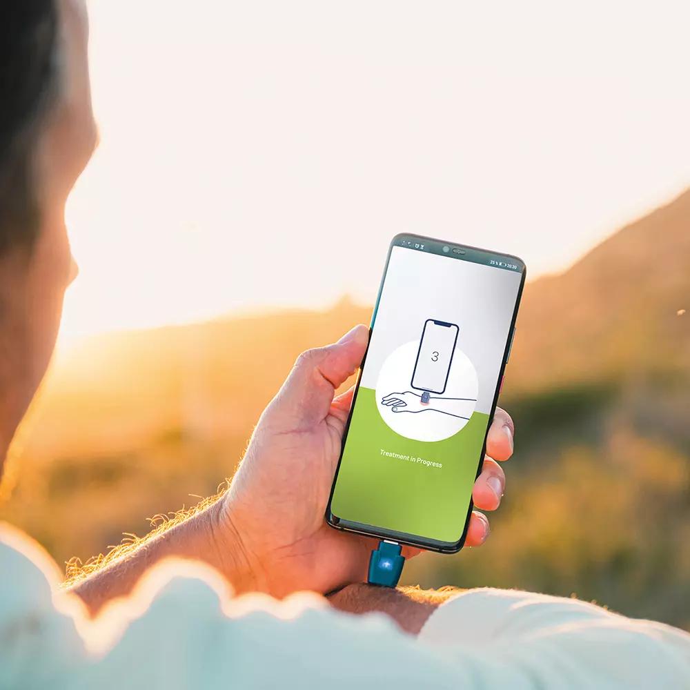 Eine Hand hält ein Smartphone mit angestecktem heat it® Insektenstichheiler, der chemiefrei bei Juckreiz/Schmerz wirkt & mit iPhone/Android kompatibel ist.