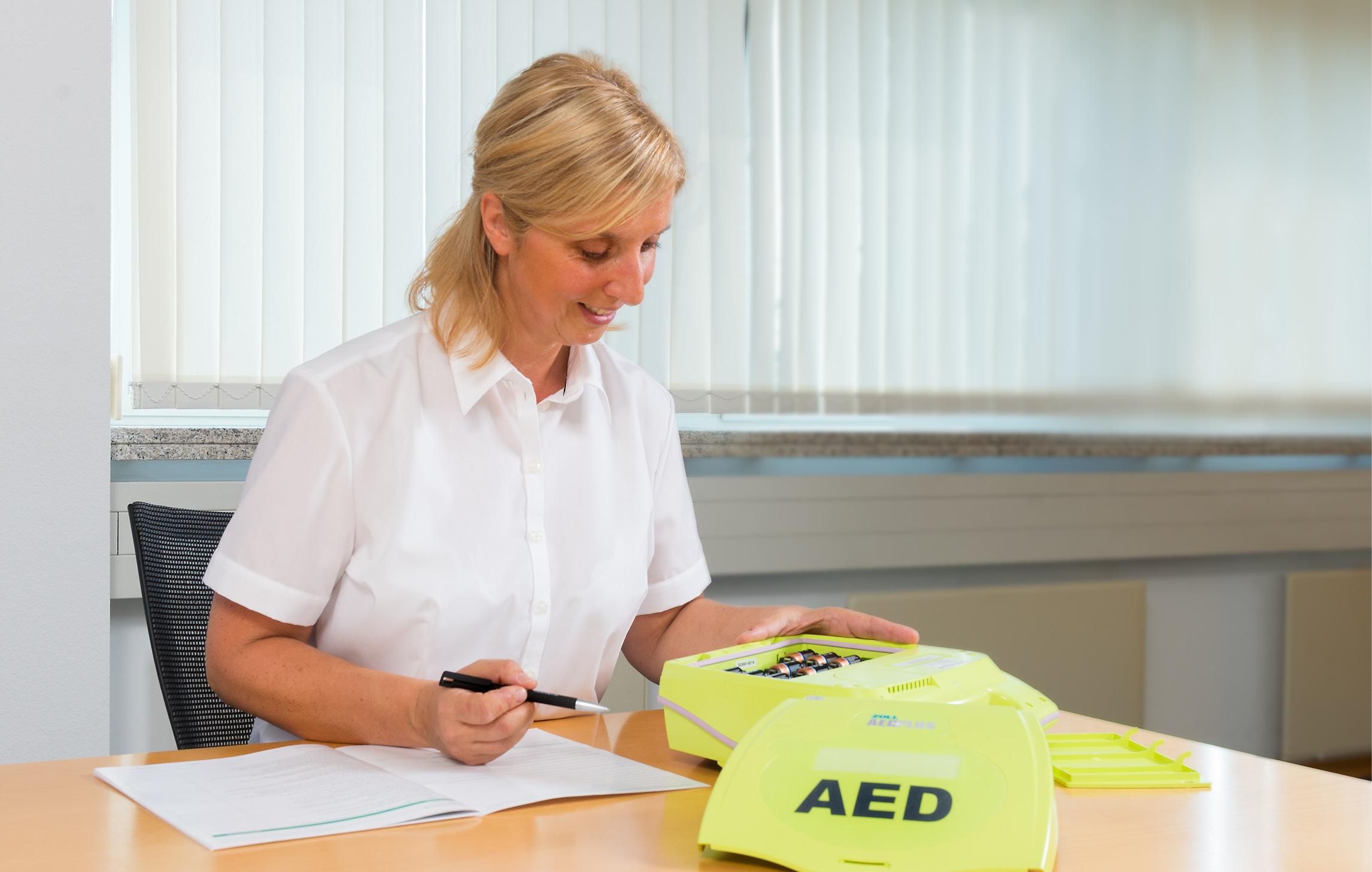 Eine Frau sitzt an einem Tisch, füllt einen Bogen Papier aus und nimmt einen neongelben AEB Defibrillator in Betrieb