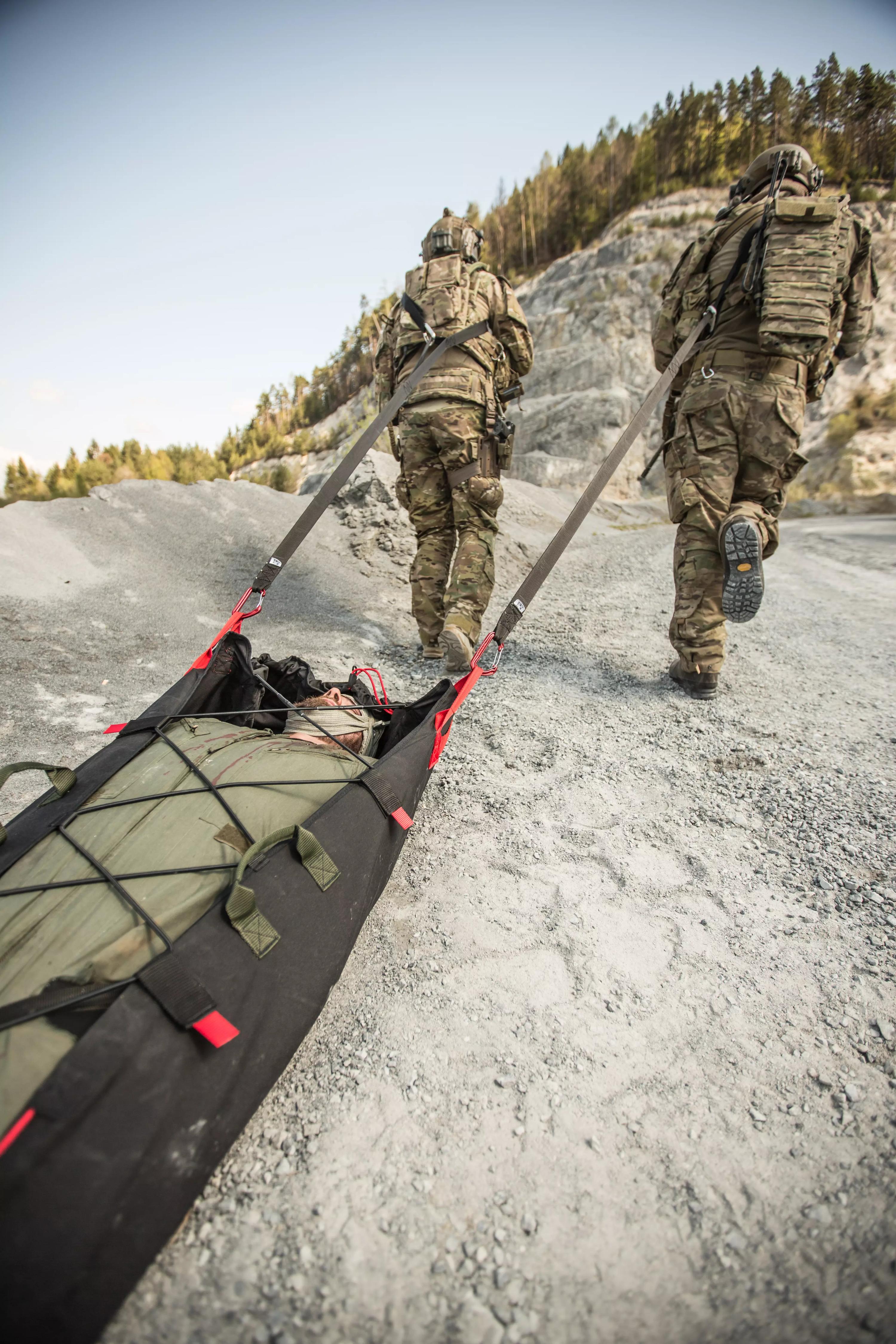 Zwei Soldaten in Tarnuniform tragen mit dem Xtract™ SR Module eine Person auf einer Trage über felsigen, spärlich bewachsenen Untergrund.