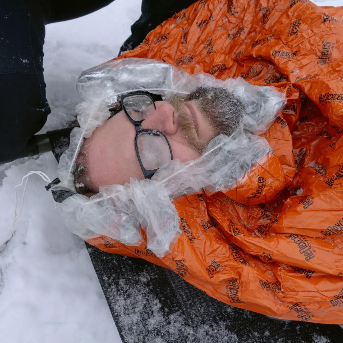 Ein Mann liegt im Schnee und ist bis zur Brust in einen Blizzard - Orange, Schlafsack mit reflektierender Blizzard Reflexcell™ Technologie eingezippt.
