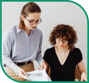 Zwei Frauen mit Brillen betrachten gemeinsam ein Blatt Papier, eine trägt ein hellblaues Oberteil, die andere ein schwarzes T-Shirt und eine Halskette.