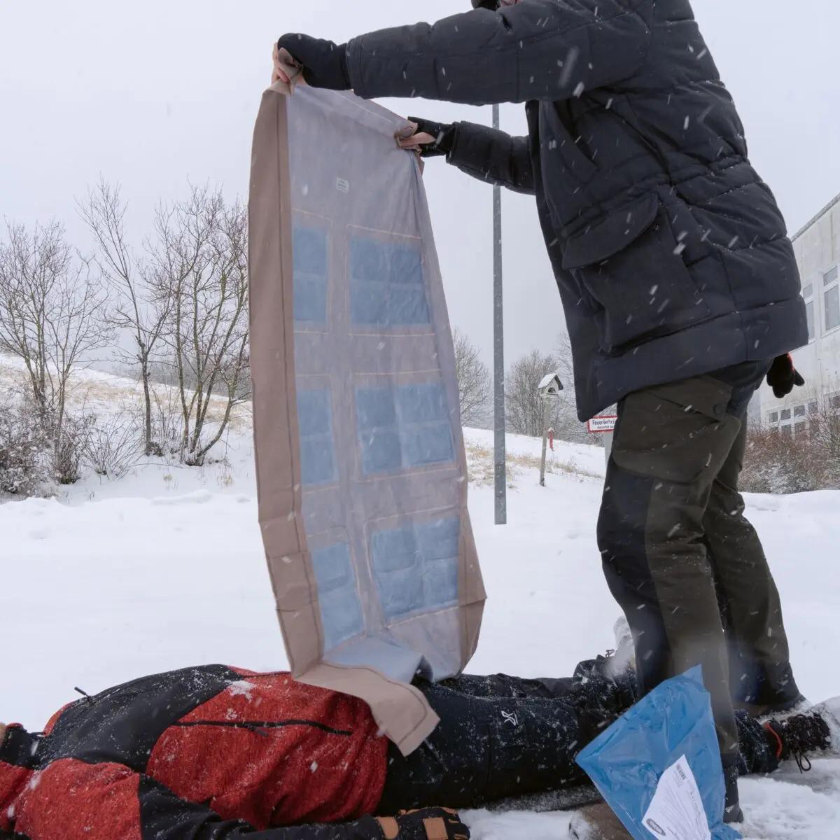 Eine Person kniet im Schnee und hilft einer anderen, die unter einer beige Ready-Heat™ - 6-Element-Decke mit luftdichter Verpackung liegt.