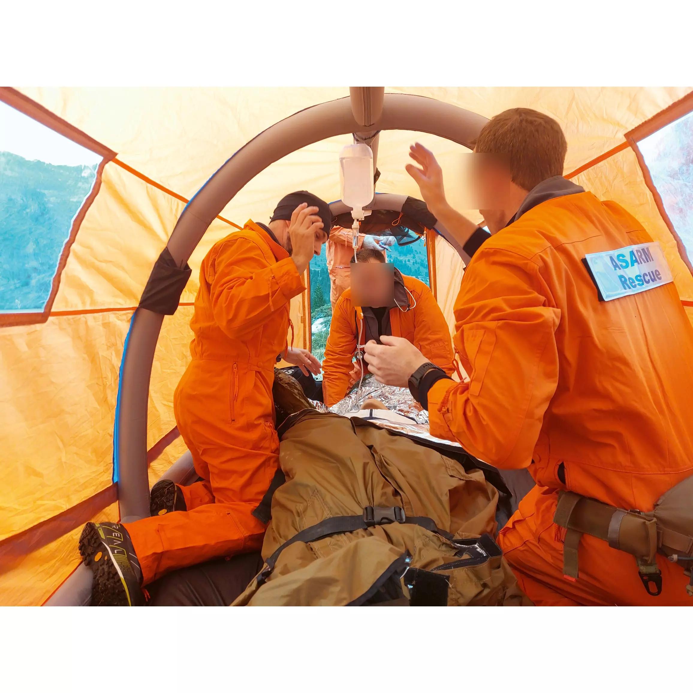 Innenansicht eines RIGLOO Rescuer Rettungszelt, in dem Rettungskräfte eine Person auf einer Trage medizinisch versorgen; Zelt leuchtend orange, leicht und sturmsicher.