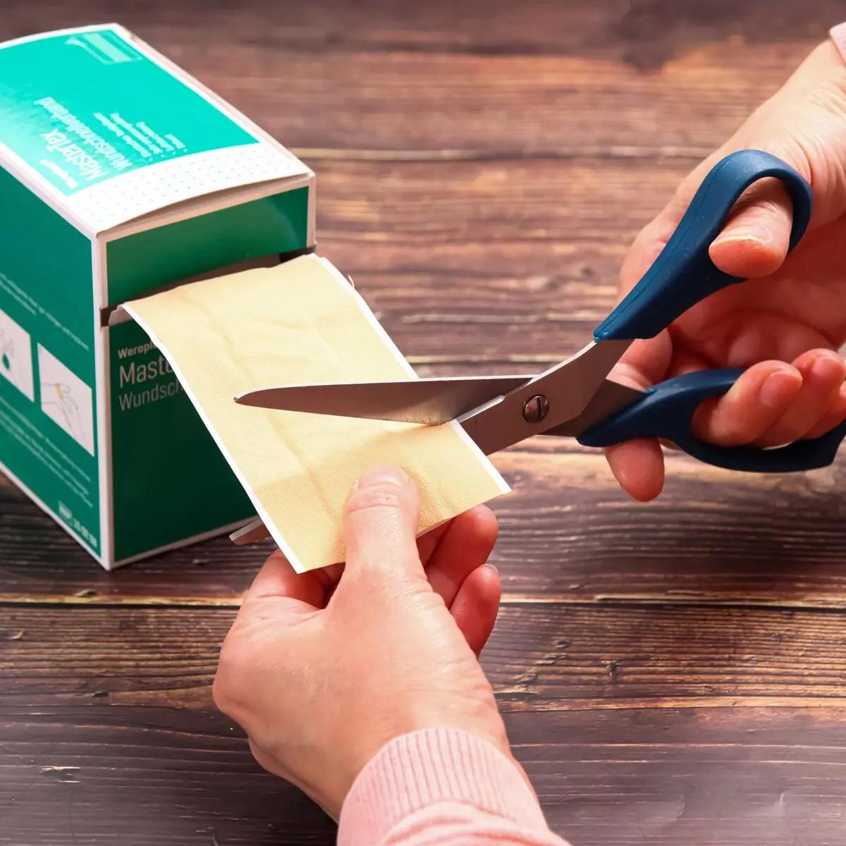 Zwei Hände schneiden mit einer blauen Schere einen Streifen des Wundschnellverband Weroplast® MasterTex von einer grünen Box mit textiler Oberfläche ab.
