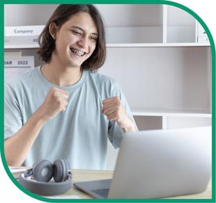 Eine Frau jubelt am Schreibtisch mit geöffnetem Laptop und grauen Over-Ear-Kopfhörern, Regal und Kalender im Hintergrund.