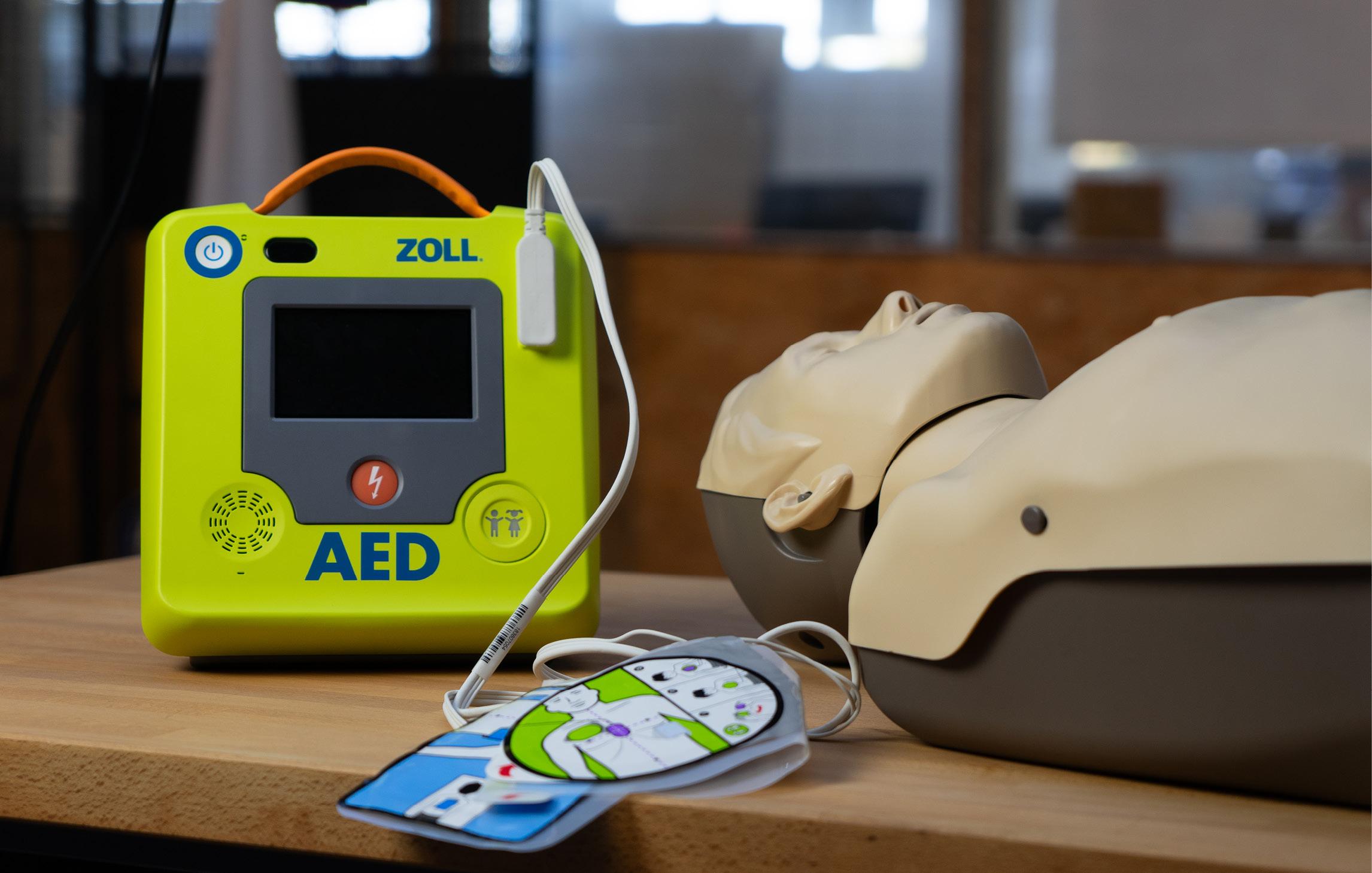 Abbildung des ZOLL AED Defibrillators mit Elektroden und der daneben liegenden Übungspuppe