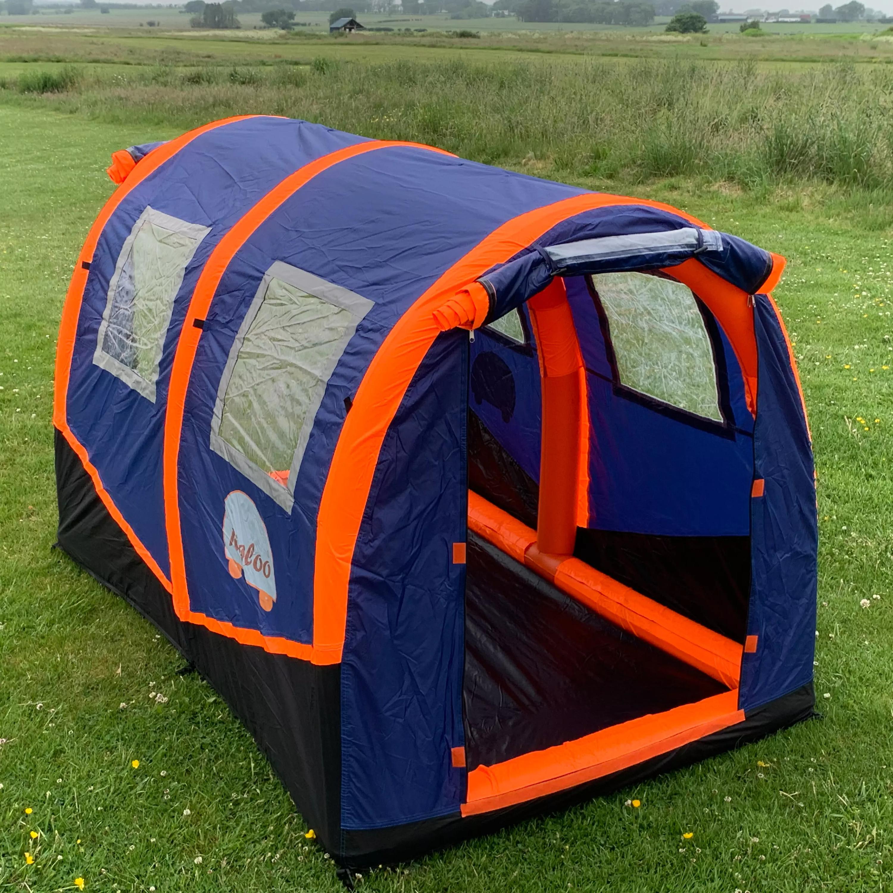 Ein blau-orangefarbenes RIGLOO Refuge Notfallzelt mit aufblasbarem Boden, gebogenem Dach, zwei seitlichen Fenstern und großer Frontöffnung steht auf einer Wiese.