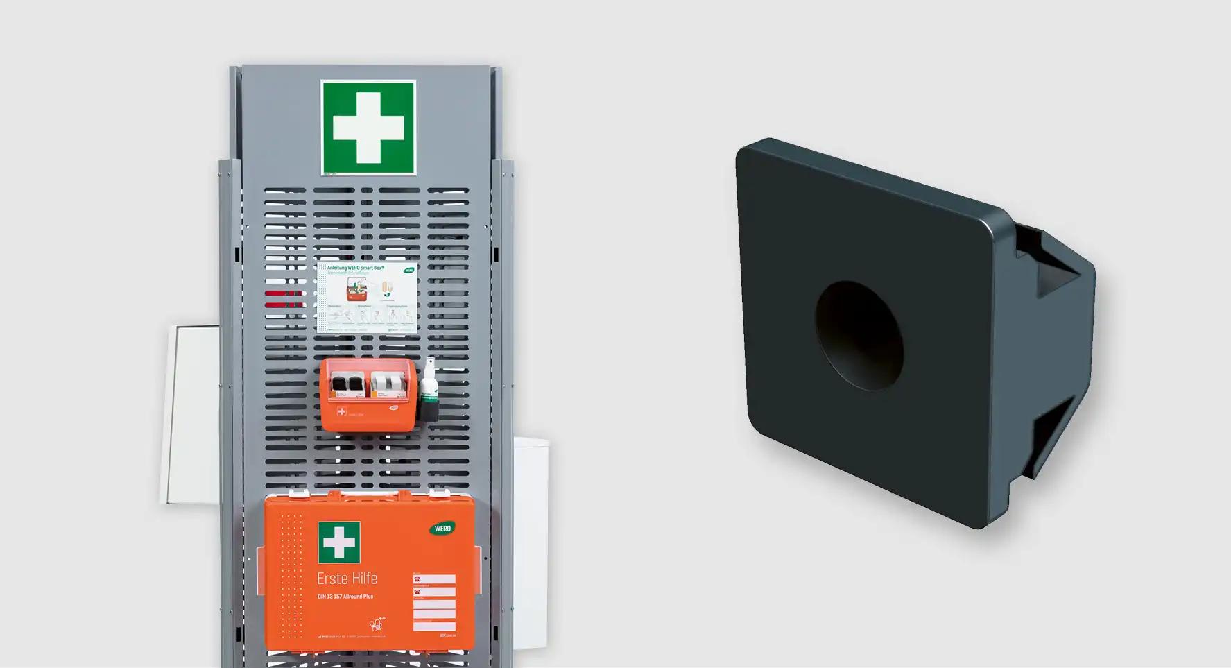 Schwarzer Notausgangs-Lockerschrank mit runder Öffnung, grünem Kreuz, Text "Erste Hilfe" und einem Zahlenschloss montiert auf einem grauen Metallrahmen.