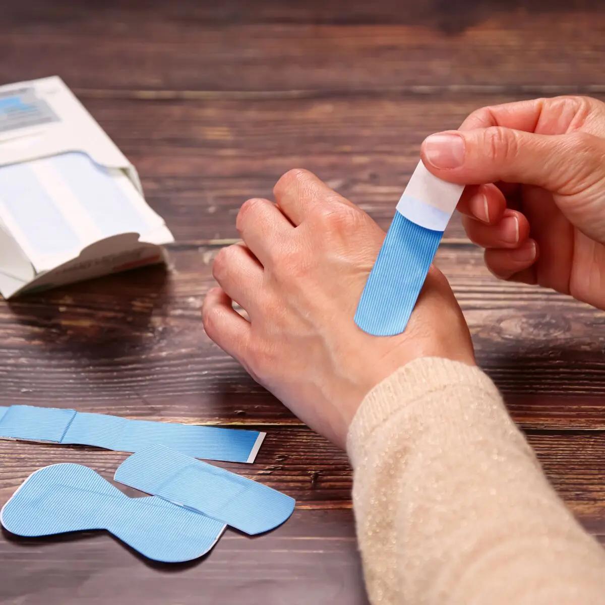 Eine Hand mit beigem Pullover hält ein blaues, detektierbares Weroplast® AquaFlex Detect Pflaster mit Aluminiumstreifen und wasserdichter Folie.