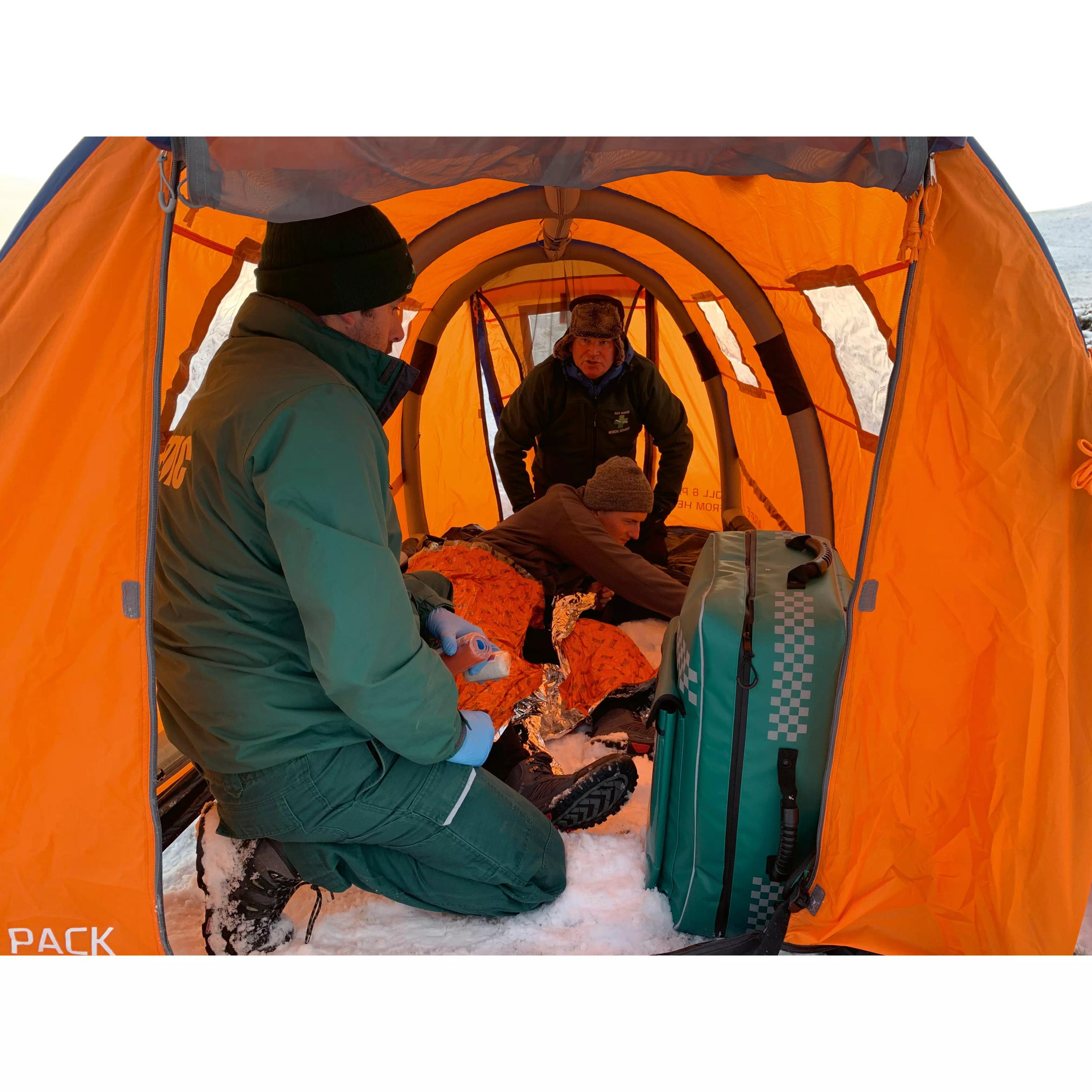 Personen mit orangefarbenen Decken leisten medizinische Hilfe in einem aufgeblasenen RIGLOO Rescuer Rettungszelt mit grüner Ausrüstung im Schnee.