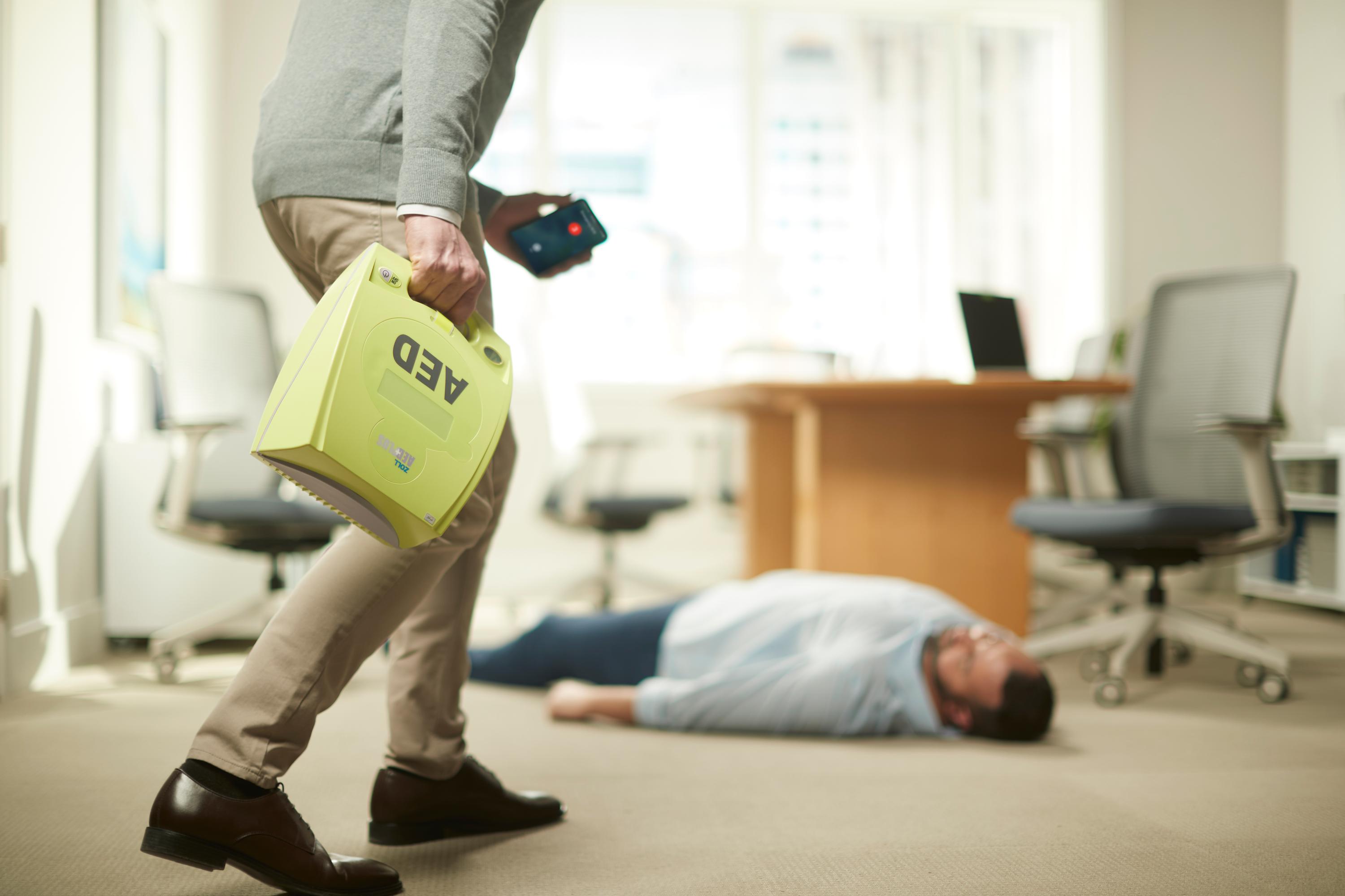 Im Büro hält eine Person liegend auf dem Boden ein rotes AED-Gerät, eine andere Person geht vorüber.