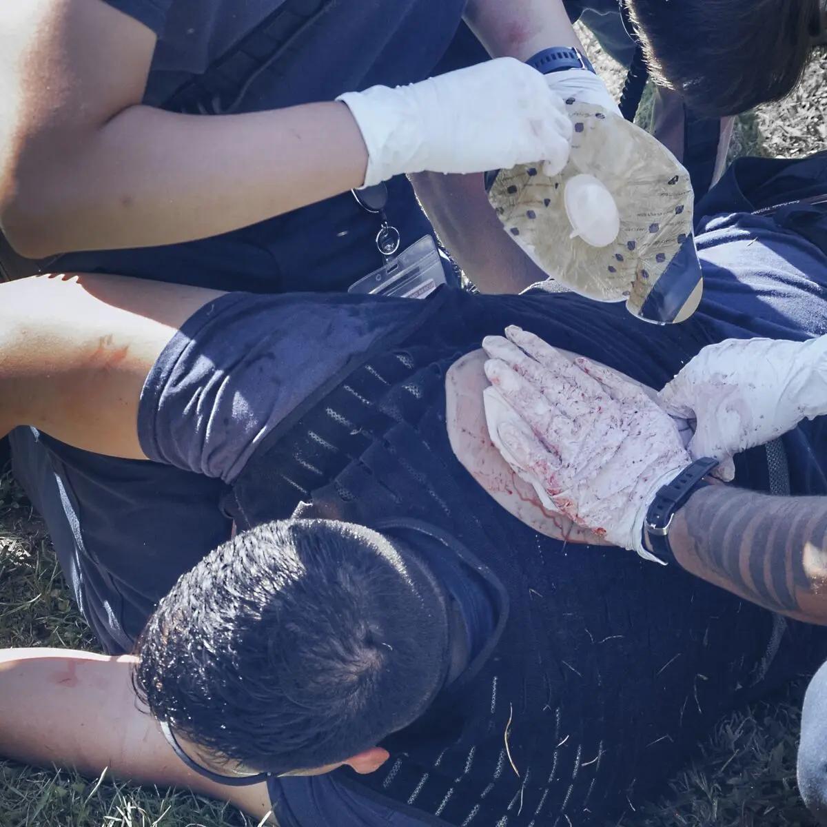 Mehrere Helfer legen am Hals einer Person im Gras mit dem TrueClot® Chest Seal Application Trainer einen Verband an, um eine Wunde realistisch zu versorgen.