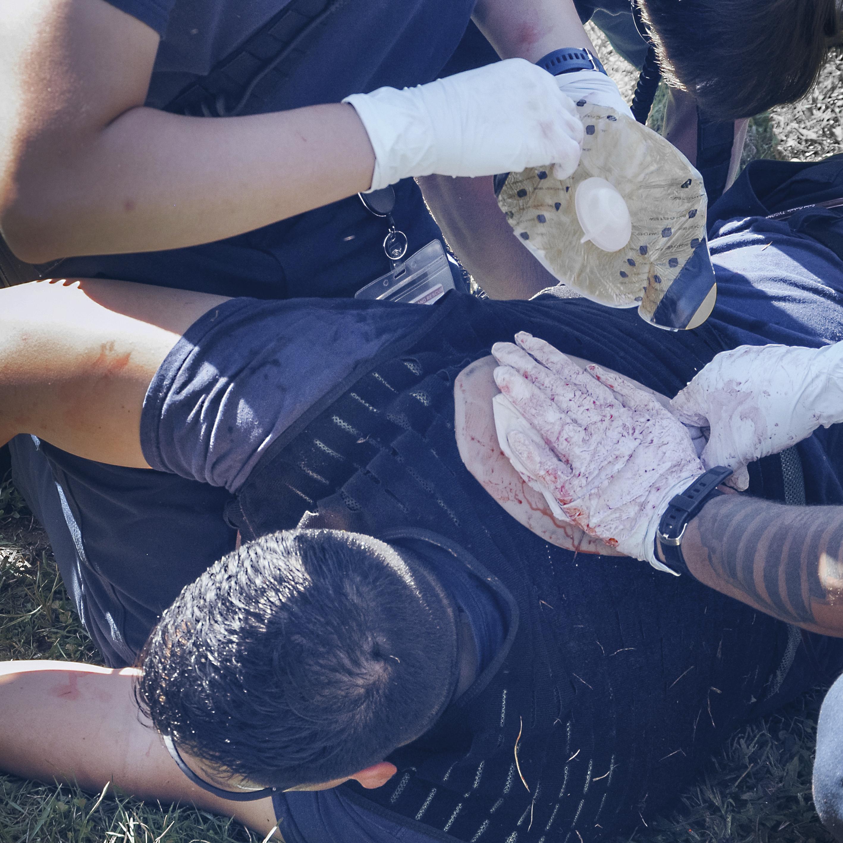 Mehrere Helfer legen am Hals einer Person im Gras mit dem TrueClot® Chest Seal Application Trainer einen Verband an, um eine Wunde realistisch zu versorgen.