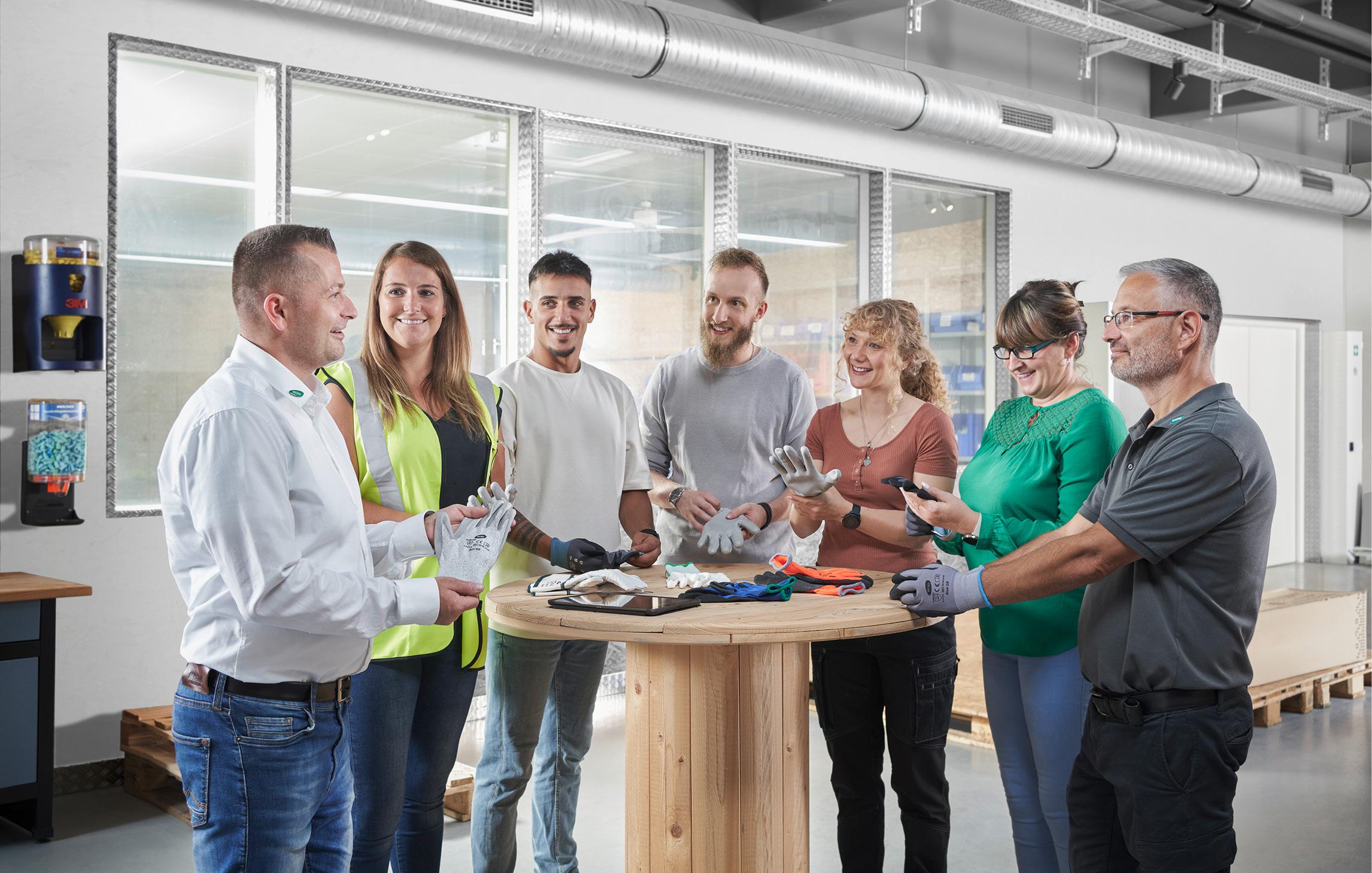 6 Mitarbeitende eines Unternehmens stehen um einen hölzernen Tisch und testen mit einem WERO Mitarbeitenden verschiedene Arbeitshandschuhe