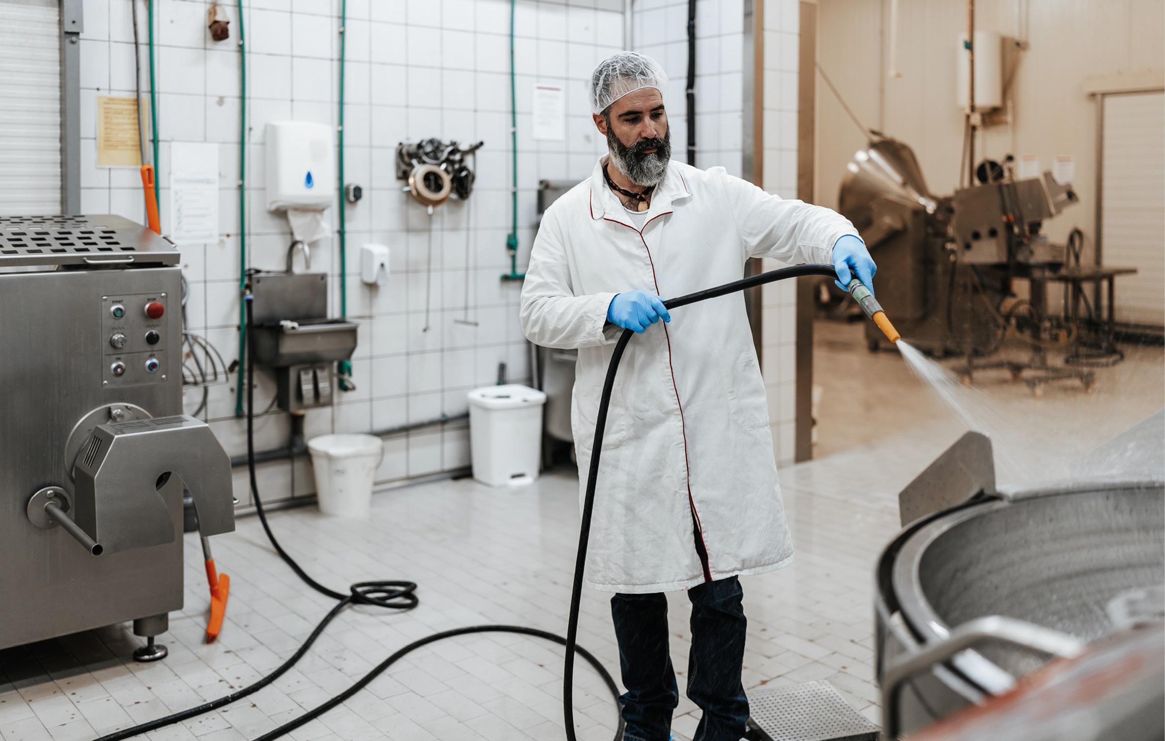 Ein Mann steht in Schutzkleidung in einem weißgekachelten Raum und wäscht mit einem Schlauch eine metallene Wanne ab