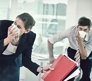 Eine Frau im schwarzen Blazer hält eine weiße Kaffeetasse ans Gesicht, neben ihr steht ein Mann mit Ordnern vor einer großen Fensterfront im Büro.