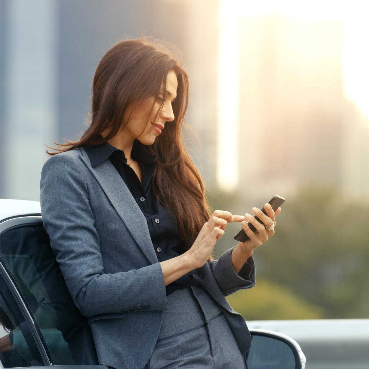 Eine Frau im grauen Anzug lehnt an einem weißen Auto und tippt konzentriert auf ihrem schwarzen Smartphone.