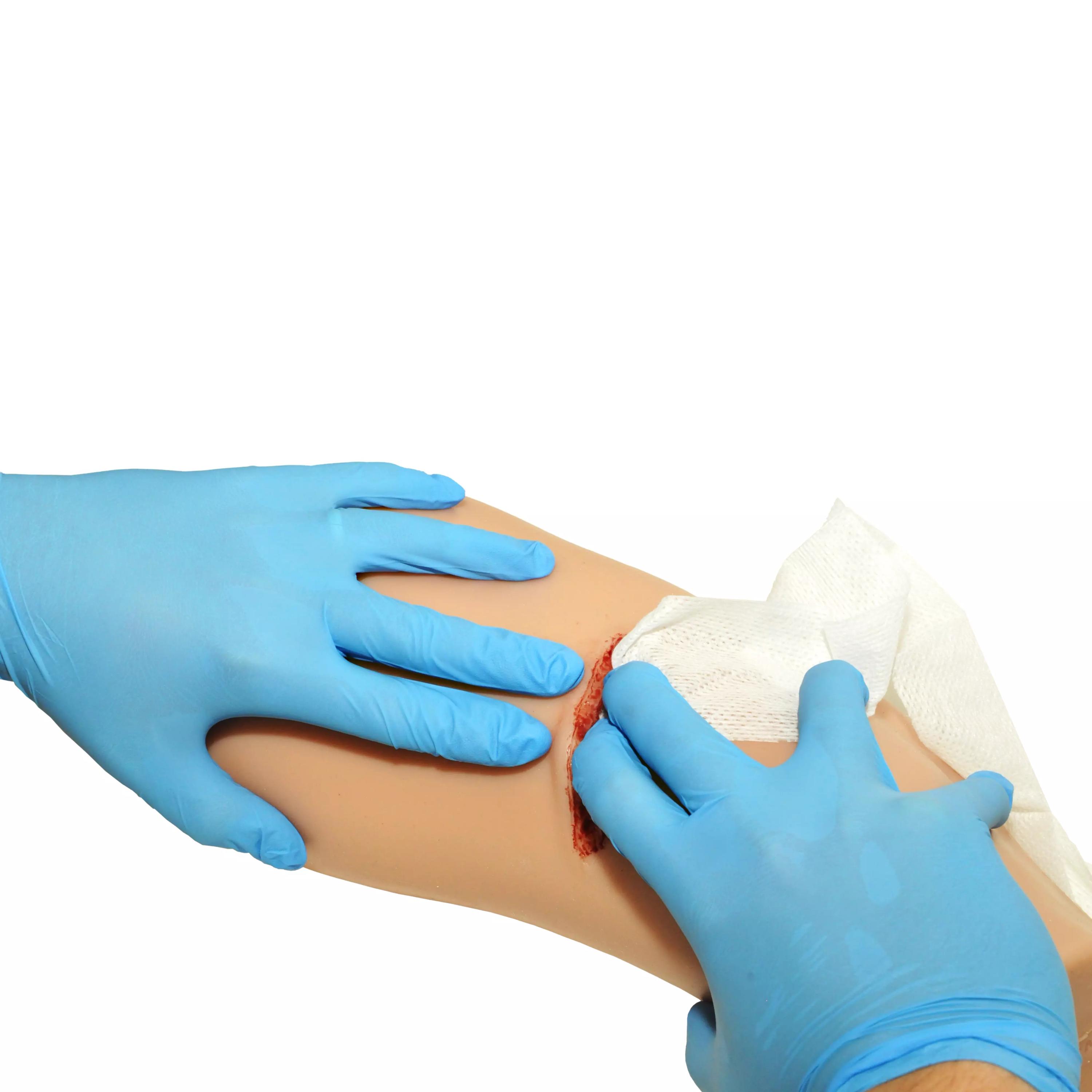 Eine blaue, behandschuhte Hand hält eine sterile Kompresse auf eine realistische Wunde am TrueClot® PCT3 Trainer mit roter Abdeckung.