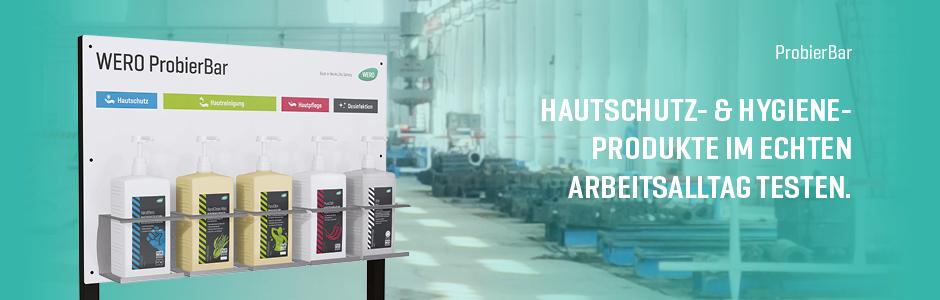 Display mit vier Flaschen Hautschutz- und Hygieneprodukten zum Testen auf einer Fabrikfläche mit Maschinen im Hintergrund.