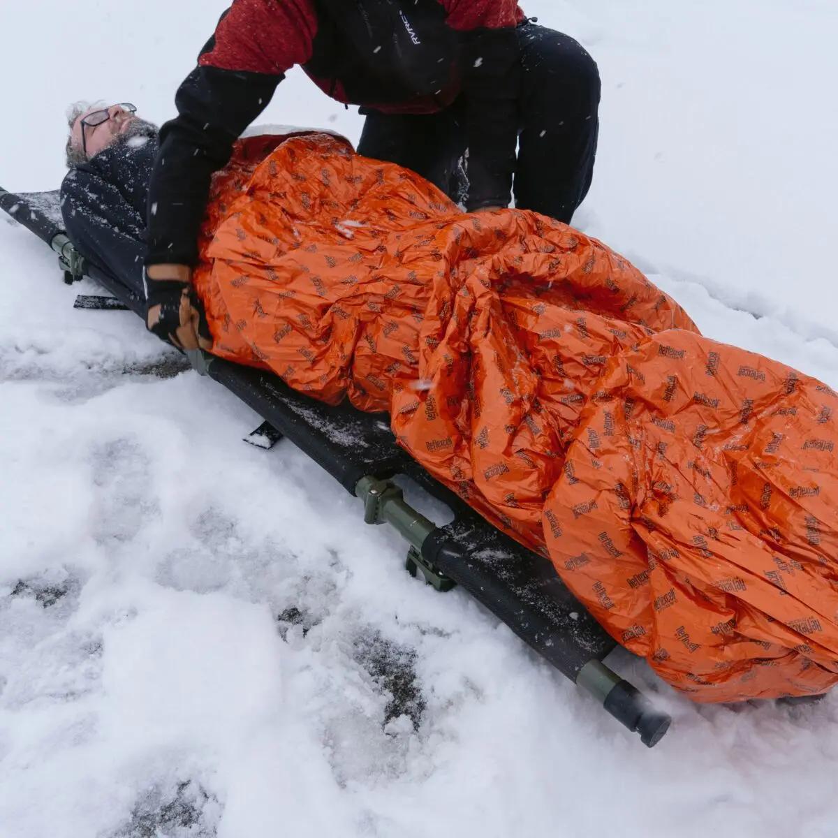 Eine Person leistet Erste Hilfe im Schnee mit einem Blizzard - Orange, Schlafsack, der einen Verletzten mit Reflexcell™ Technologie wärmt und vor Kälte schützt.