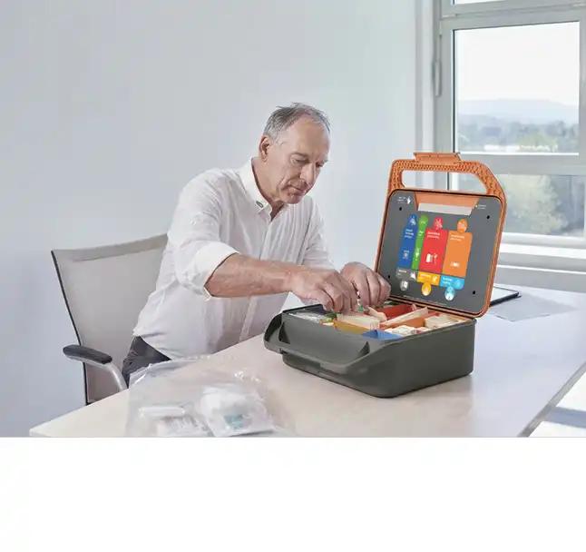 Ein älterer Mann sitzt am Tisch und bedient eine geöffnete orange-graue Werkzeugbox mit Bildschirm und bunten Bedientasten.