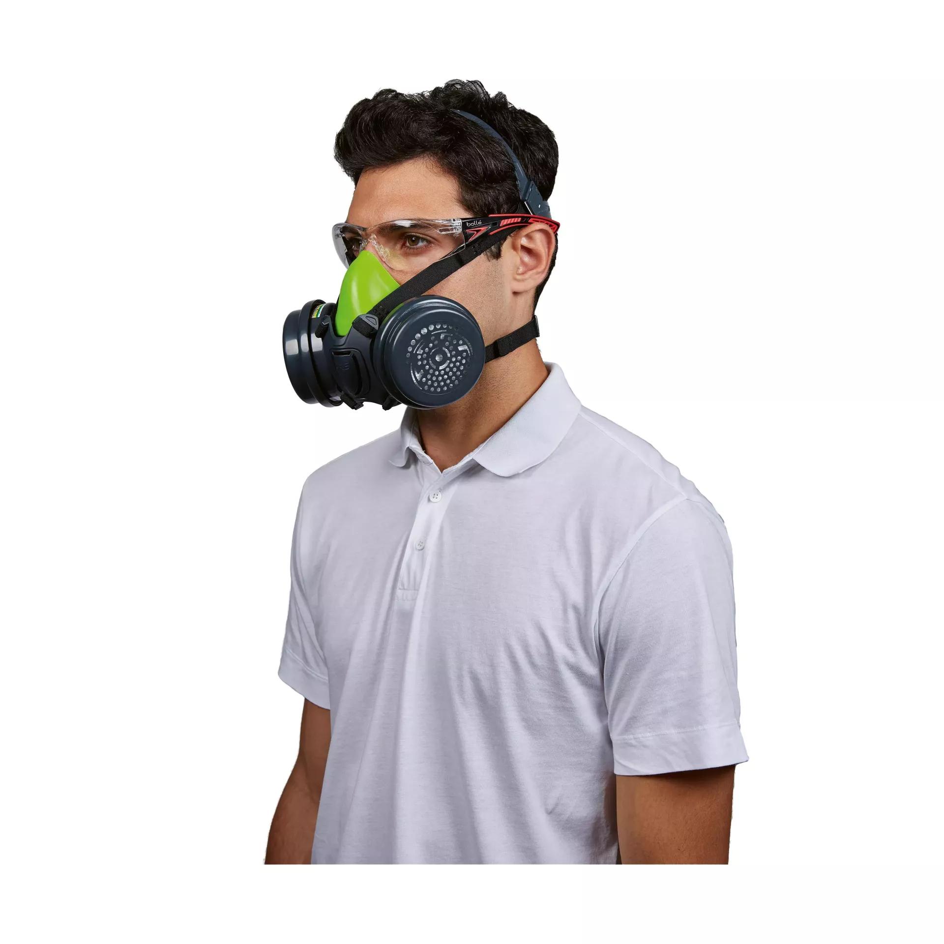Ein Mann trägt eine schwarze Gasmaske mit grünem Filter und klare Schutzbrille, dazu ein weißes Polohemd.