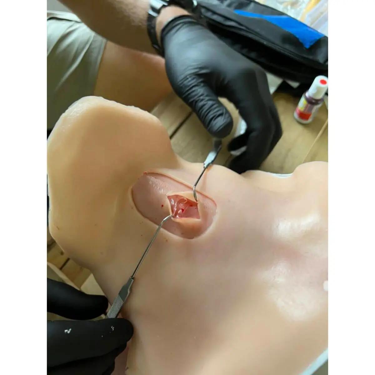 Ein schwarzhandschuhter Arm nutzt Metallscheren am Battlefield Partners Desktop CRIC Trainer, um präzise einen Schnitt an der Hinterkopfhaut des Modells zu üben.