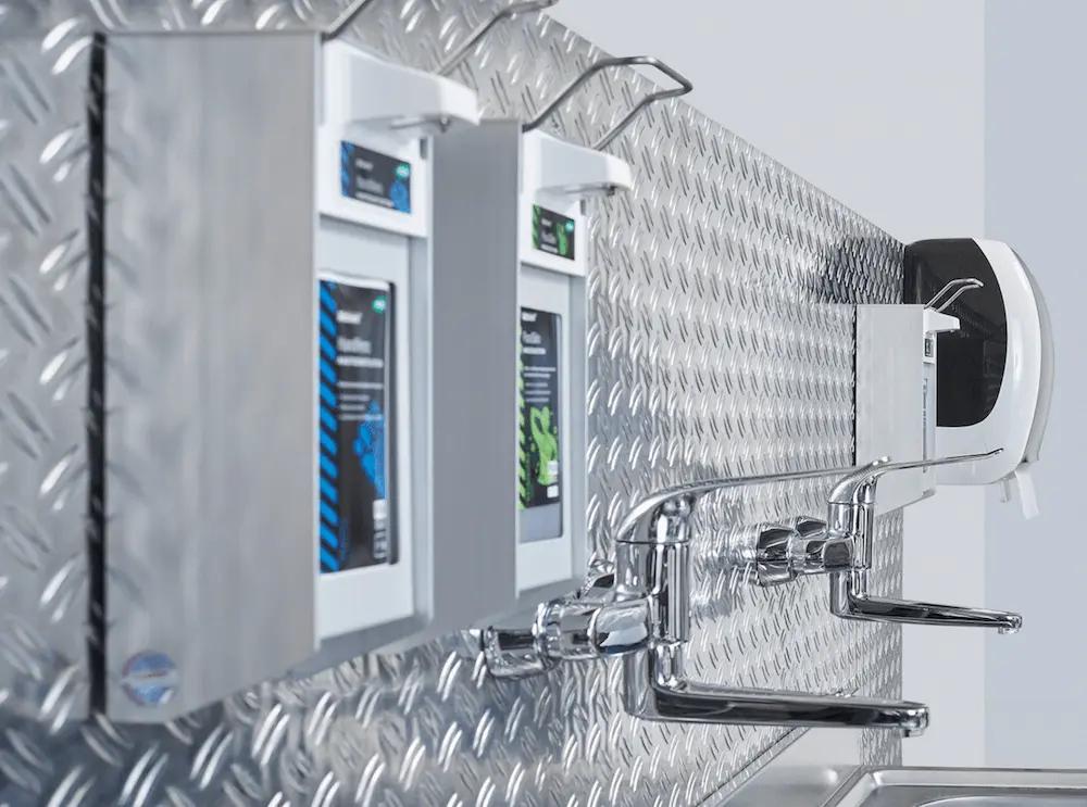 Zwei Edelstahl-Handwaschstationen mit weißen Spendern und Chromarmaturen sind an einer Wand mit Rautenmuster montiert.