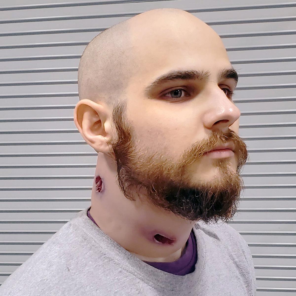 Ein Mann trägt eine Techline Moulage Schusswunde Nacken, die als simulierte, durchgehende Nacken-Schussverletzung mit sichtbarem Gewebe gestaltet ist.