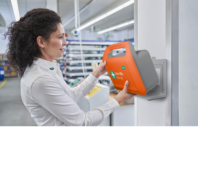 Frau mit lockigen Haaren bedient ein orange-graues Wandgerät mit Griff und grünem Logo in industrieller Umgebung.