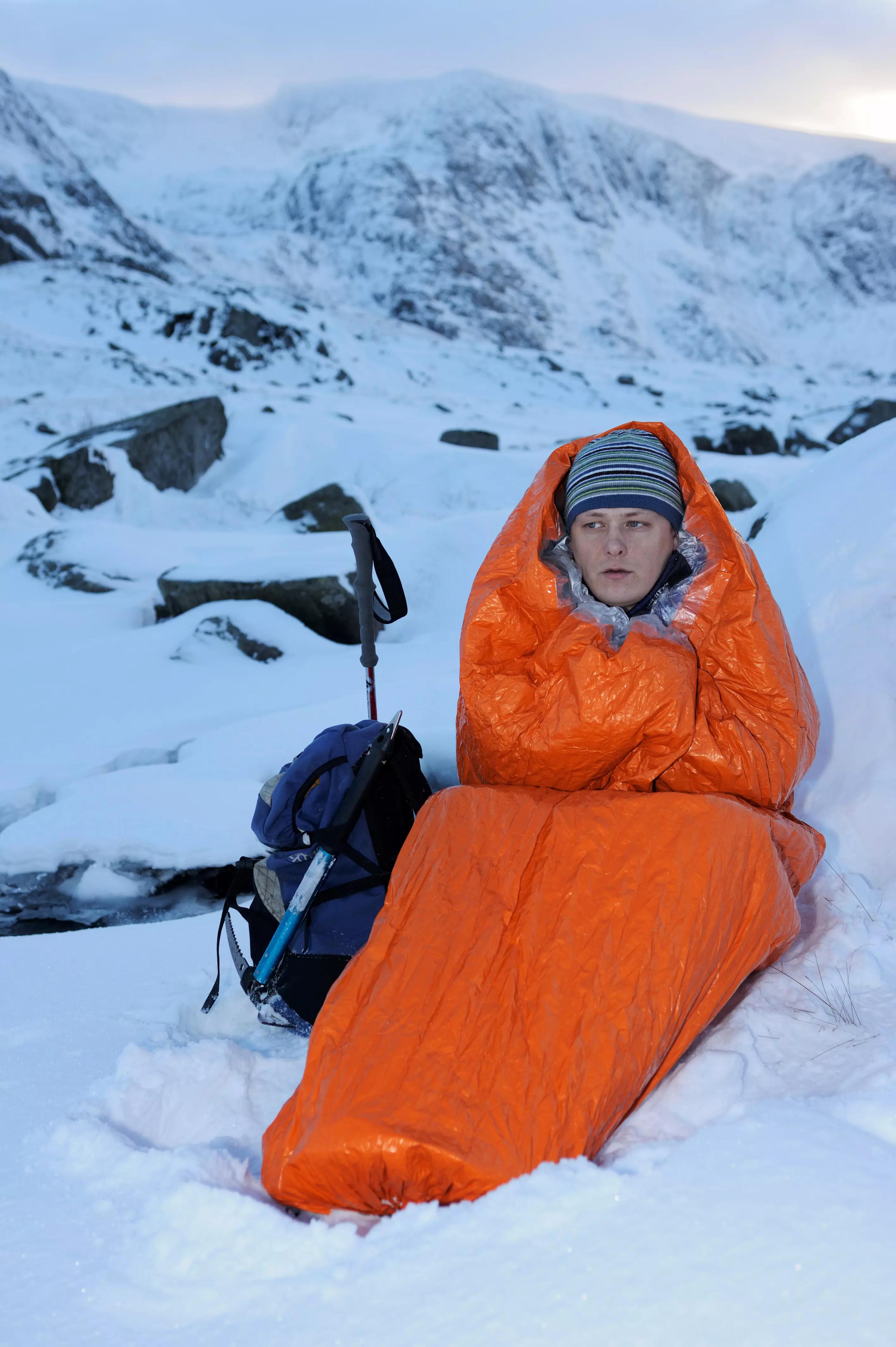 Eine Person liegt im Blizzard - Orange, Schlafsack auf Schnee, mit sichtbar gesteppter Struktur und Blizzard Reflexcell™ Technologie, umgeben von Wanderzubehör.