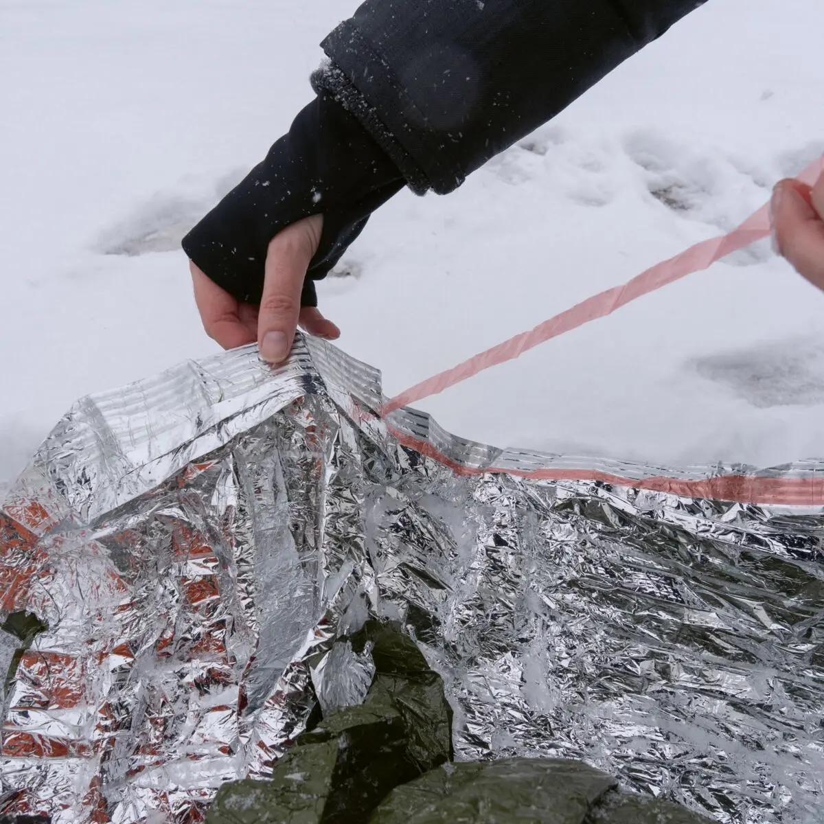 Eine Hand hält eine Blizzard - Grün, Decke mit reflektierendem, mehrlagigem Aluminium-Verbundmaterial und zellartiger Struktur im Schnee.
