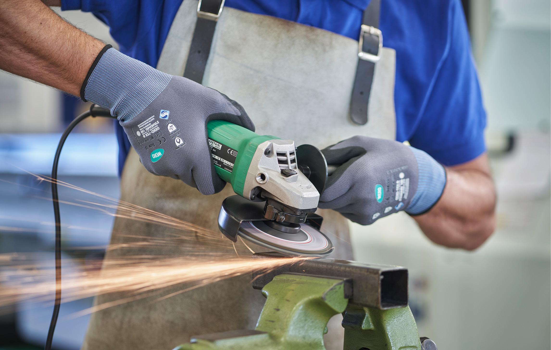 Ein Mann steht mit lederner Schürze, WERO Arbeitshandschuhen und Flex vor einem aufgebockten Metalllstück. Funkenflug