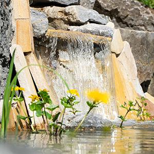 Mehrstufiger Steingarten-Wasserfall mit fließendem Wasser in einen Teich, umgeben von gelben Blumen und grüner Bepflanzung