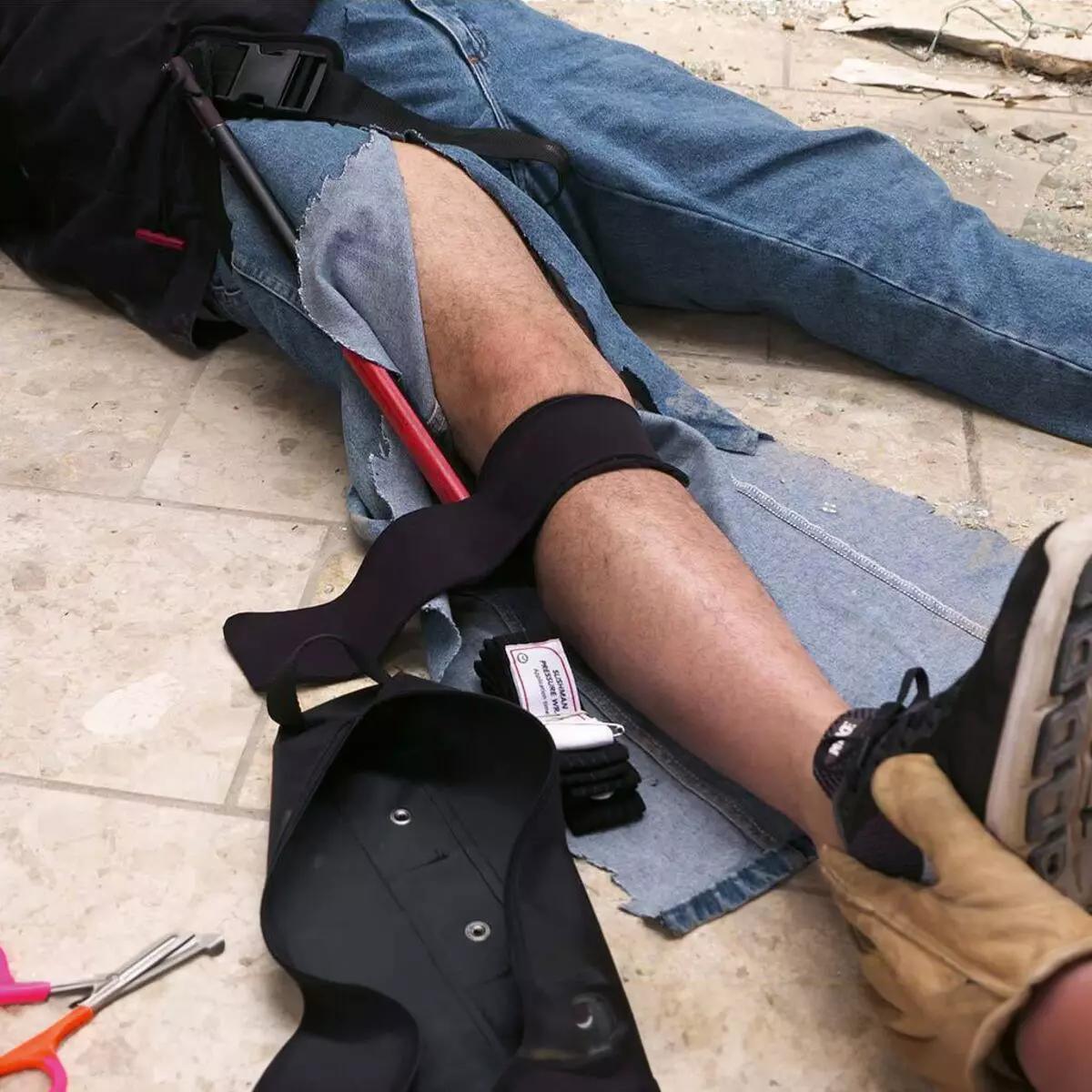 Eine Person erhält Notfallhilfe mit angelegter Slishman Traction Splint - COMPACT (STS-C) am Bein, schwarzer Gurt und roter Stab sichern das verletzte Bein.