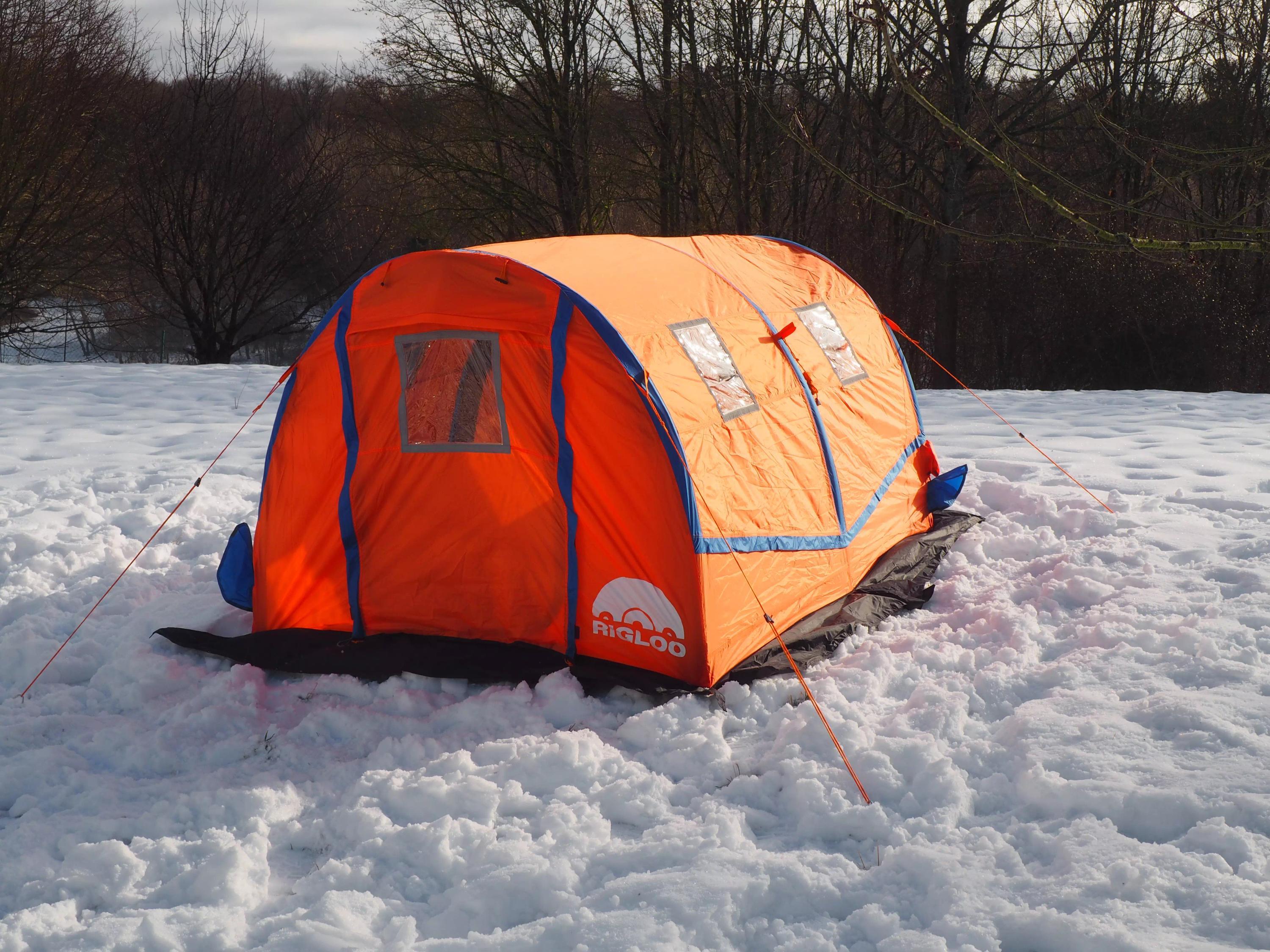 Ein orange-blaues RIGLOO Rescuer Rettungszelt mit stabilem Gestänge, zwei seitlichen Fenstern und abnehmbarer Bodenplane steht im verschneiten Wald.