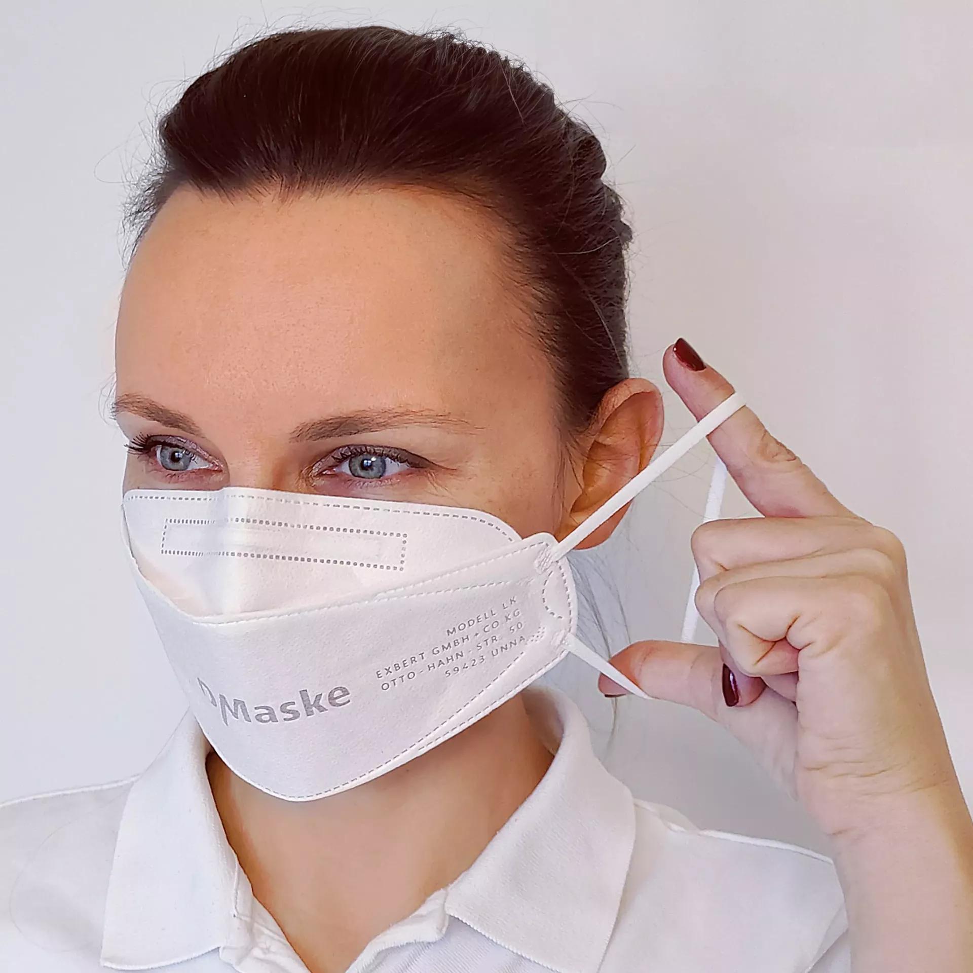 Frau mit weißem Hemd trägt eine weiße Schutzmaske mit schwarzer Beschriftung „Expert Mask Model 1K GBT-VERN Maske“, dunkle Haare, lackierte Nägel.