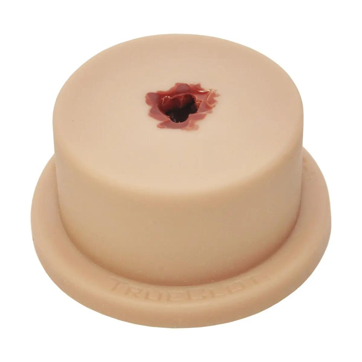 Runder TrueClot® Basic Packing Trainer mit beiger Oberfläche, zentralem Loch mit dunkelroter Flüssigkeit und flacher Standbasis mit TRUEGLOT-Prägung.